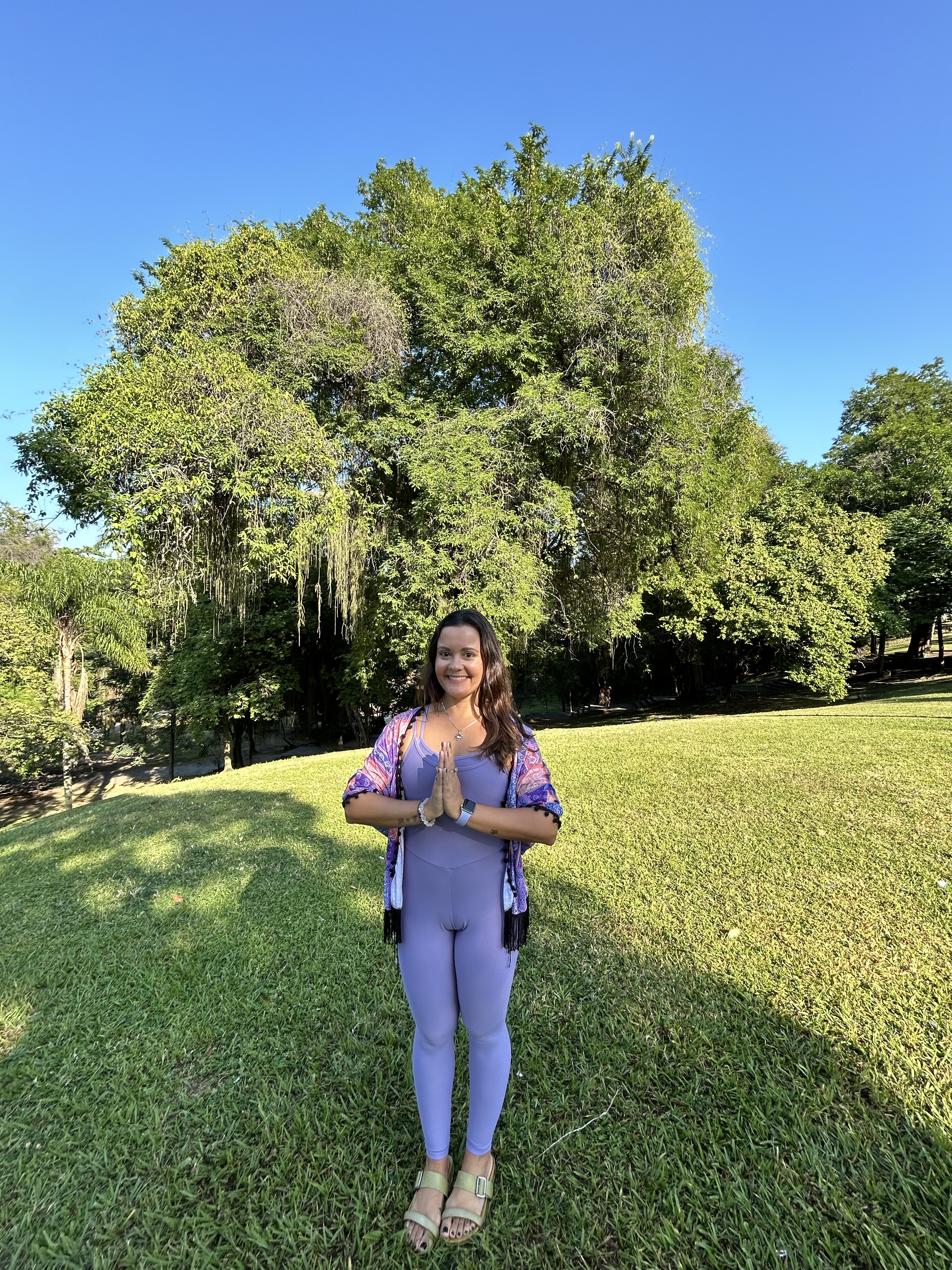 Dani Allexándre em Anjali Mudra, com as mãos unidas em gesto de oração, sorrindo em meio à natureza